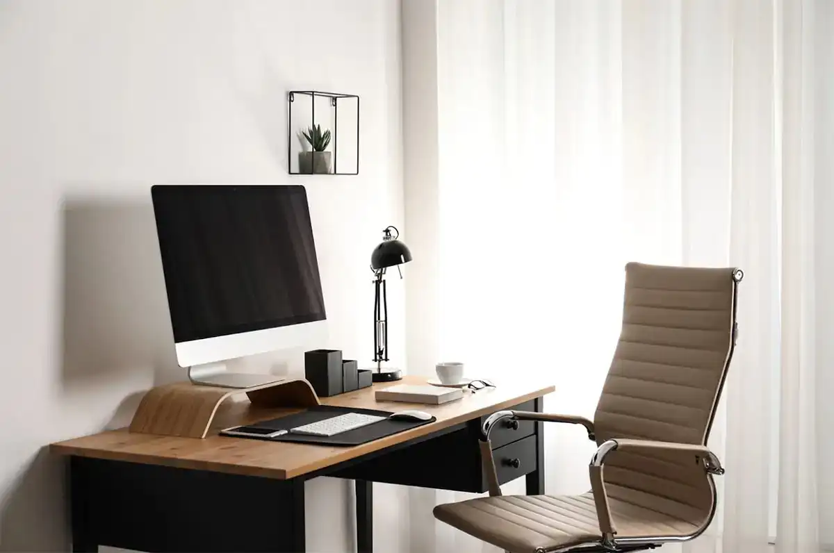 Minimalist home office with a computer, keyboard, mouse, lamp, cup, and notepad on a wooden desk. A tan swivel chair is next to the desk, and sheer curtains cover the window. A small plant is on a wall shelf.